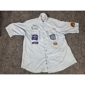 May include: A light blue short-sleeve button-down shirt featuring multiple embroidered patches and text including Missouri Bass Federation 2004 State Team Jim White, Lowrance, and Pure Fishing. The shirt has two front chest pockets and a collar.