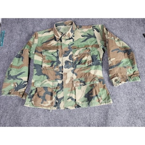 May include: Camouflage pattern jacket with four front pockets and buttoned cuffs. The jacket features a collared neckline and a button-up front. The color scheme includes shades of green, brown, and tan, creating a military-inspired aesthetic.