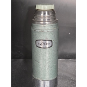May include: A vintage green, textured metal vacuum flask with a silver-coloured base and neck. The bottle features a black and silver label with the text Aladdin's STANLEY thermos. It is topped with a light-coloured, screw-on plastic cap.