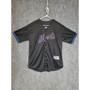 Puede incluir: Camiseta de béisbol negra con la palabra "Mets" en escritura naranja y azul en el pecho. La camiseta tiene una parte delantera abotonada, mangas cortas con ribete azul y una pequeña etiqueta en la parte inferior derecha.