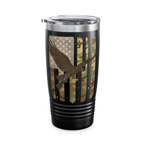 Op de afbeelding: Zwarte geïsoleerde beker met een zilveren deksel. De beker heeft een versleten Amerikaans vlagontwerp met een camouflagepatroon en een bruine adelaar. De beker is ontworpen om dranken warm of koud te houden.