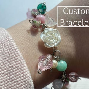 May include: A custom bracelet with a variety of charms, including a white rose, pink and green beads, and a clear crystal. The bracelet is on a person's wrist and the text "Custom Bracelets" is visible in the image.