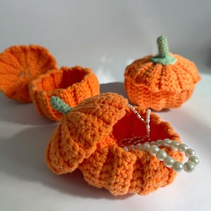 Puede incluir: Cuatro contenedores de calabaza de ganchillo, uno con la tapa abierta que revela un collar de perlas en el interior. Los contenedores son de color naranja y tienen tallos verdes.