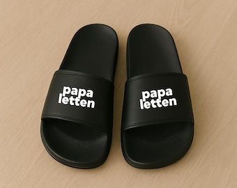 Könnte beinhalten: Ein Paar schwarze Badesandalen mit dem weißen Schriftzug "papa letten" auf der Oberseite. Die Sandalen sind aus glattem Material gefertigt und werden auf einer hellbraunen Oberfläche präsentiert.