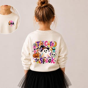 Könnte beinhalten: Ein cremefarbenes Sweatshirt mit einer bunten Halloween-Grafik auf der Rückseite, die einen Geist mit Hexenhut, einen Kürbis, einen Totenkopf und den Text Too Cute to Spook zeigt. Auf der Brust steht der Name LINA in bunten Buchstaben.