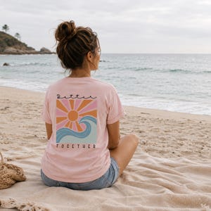 Peut inclure: Un t-shirt rose clair &agrave; manches courtes avec un motif au dos. Le design repr&eacute;sente un soleil stylis&eacute; aux rayons orange et roses au-dessus d'une vague bleue. Le texte "Better" est imprim&eacute; au-dessus du soleil et "TOGETHER" sous la vague.