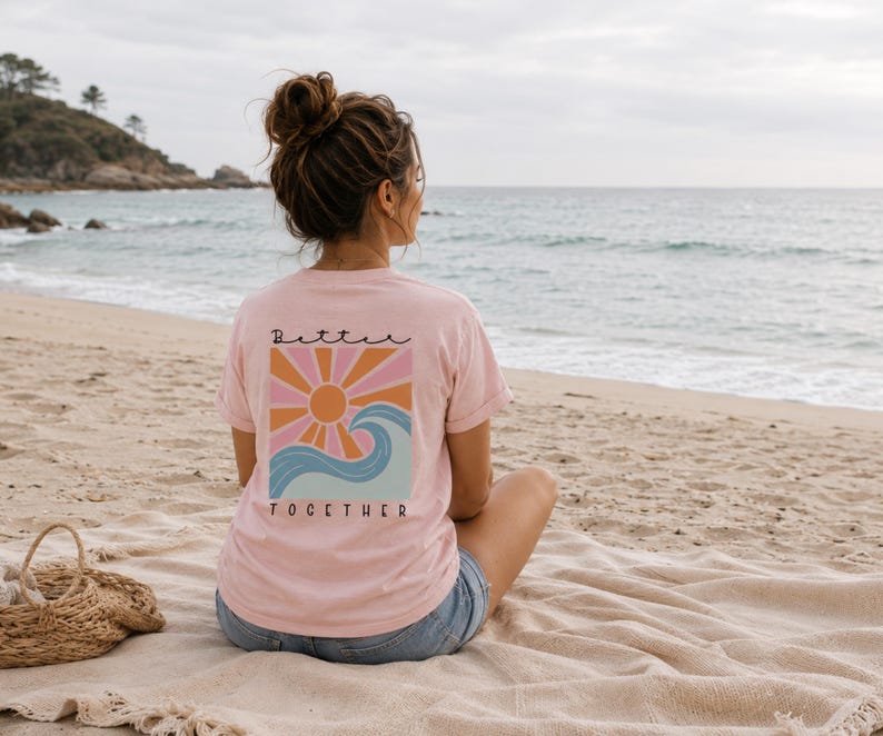 Peut inclure: Un t-shirt rose clair &agrave; manches courtes avec un motif au dos. Le design repr&eacute;sente un soleil stylis&eacute; aux rayons orange et roses au-dessus d'une vague bleue. Le texte "Better" est imprim&eacute; au-dessus du soleil et "TOGETHER" sous la vague.