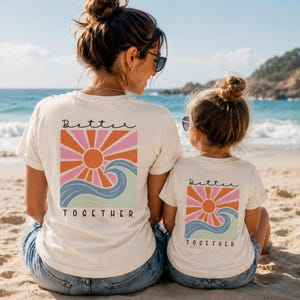 Peut inclure: Deux t-shirts blanc cass&eacute; avec un motif au dos repr&eacute;sentant un soleil orange stylis&eacute;, des rayons roses et une vague bleue. Le texte "Better TOGETHER" est imprim&eacute; en noir au-dessus et en dessous du motif, cr&eacute;ant un ensemble assorti.
