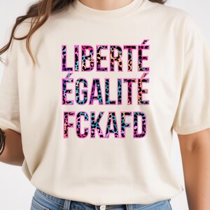 Pode incluir: Uma t-shirt de manga curta na cor creme com o texto LIBERTÉ ÉGALITÉ FCKAFD impresso num padrão de leopardo multicolorido com tons de rosa, azul e amarelo. As letras têm um contorno rosa forte sobre o tecido claro.