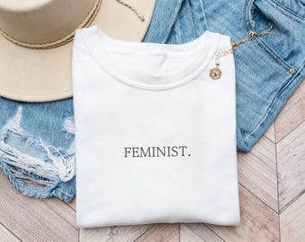 May include: White t-shirt with the word "FEMINIST." printed in black on the front. The shirt is folded and laid on a denim background.