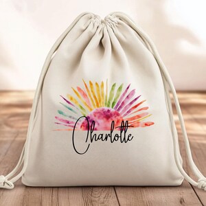 Può includere: Una borsa a cordoncino bianca con un design di alba acquerellata colorata e il nome "Charlotte" stampato sotto.