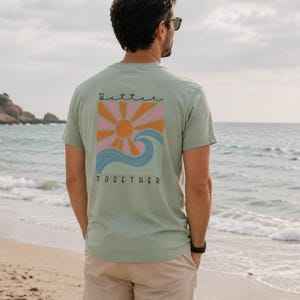 Peut inclure: Un t-shirt &agrave; manches courtes vert sauge avec un motif au dos. Le design pr&eacute;sente un soleil orange stylis&eacute; avec des rayons roses et orange au-dessus d'une vague bleue. Le texte "Better" est &eacute;crit en noir au-dessus et "TOGETHER" en dessous.