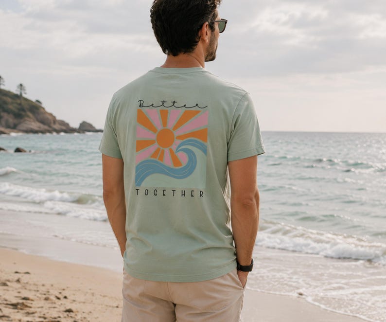 Peut inclure: Un t-shirt &agrave; manches courtes vert sauge avec un motif au dos. Le design pr&eacute;sente un soleil orange stylis&eacute; avec des rayons roses et orange au-dessus d'une vague bleue. Le texte "Better" est &eacute;crit en noir au-dessus et "TOGETHER" en dessous.