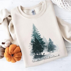 Könnte beinhalten: Cremefarbenes Sweatshirt mit einem Walddruck, der immergrüne Bäume und eine Hirschsilhouette zeigt. Die Worte "Winter Wonderland" sind in Schreibschrift geschrieben. Dekorative Kürbisse sind in der Nähe platziert.