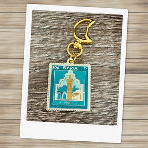 Könnte beinhalten: Goldfarbener Schlüsselanhänger mit einem blauen und weißen Emaille-Stempel mit dem Text "20f SYRIA Y." und einer Darstellung einer Moschee.