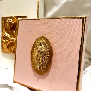 May include: A square canvas painted in light pink with a gold-painted edge, featuring a centered oval gold-toned relief plaque of the Virgin of Guadalupe. A second white canvas with a similar gold-toned relief plaque is visible in the background.