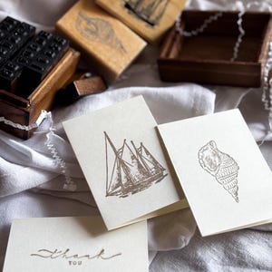 May include: A collection of nautical-themed stamps and stationery. The image shows wooden stamps with sailboat and seashell designs, along with blank cards and a card that says "thank you". The items are arranged on a white cloth.