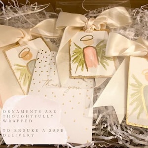 May include: Rectangular ornaments featuring hand-painted angel illustrations with gold leaf edges, each tied with a cream satin ribbon. A white card with gold polka dots and the text "Thank you" is included, alongside a note stating "Ornaments are thoughtfully wrapped to ensure a safe delivery."