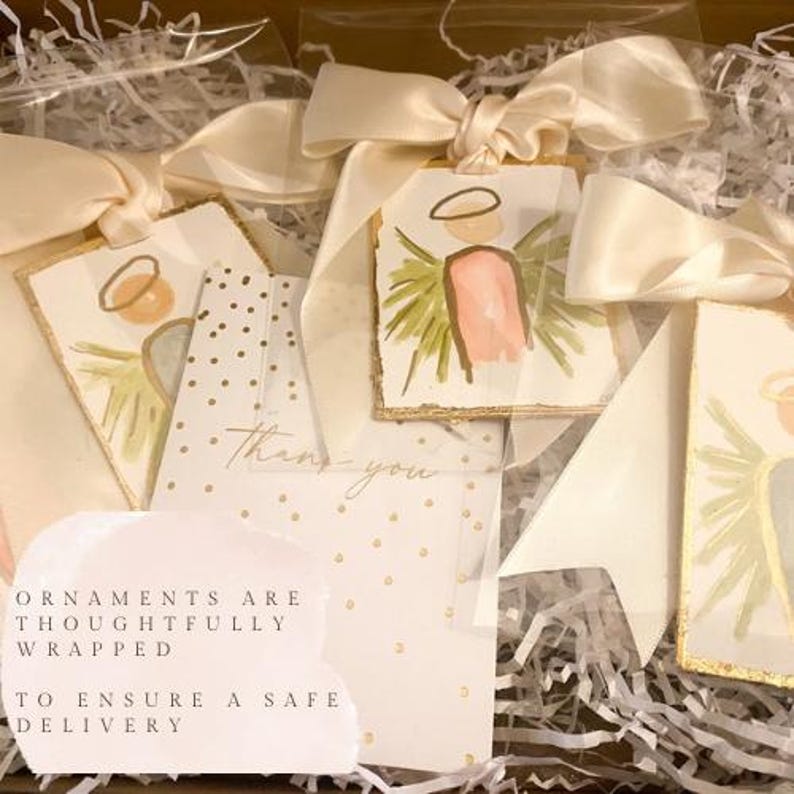 May include: Rectangular ornaments featuring hand-painted angel illustrations with gold leaf edges, each tied with a cream satin ribbon. A white card with gold polka dots and the text "Thank you" is included, alongside a note stating "Ornaments are thoughtfully wrapped to ensure a safe delivery."