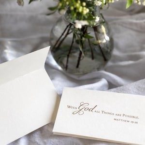 May include: Ivory-colored greeting cards with the text "With God, All Things Are Possible. Matthew 19:26" in a script font. One card is open, revealing a blank interior. A glass vase with white flowers is in the background.