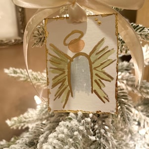 May include: A rectangular paper ornament featuring a hand-painted angel with a gold halo, light blue body, and gold-toned wings. The edges are accented with gold leaf, and the piece is suspended by a cream-colored satin ribbon bow.