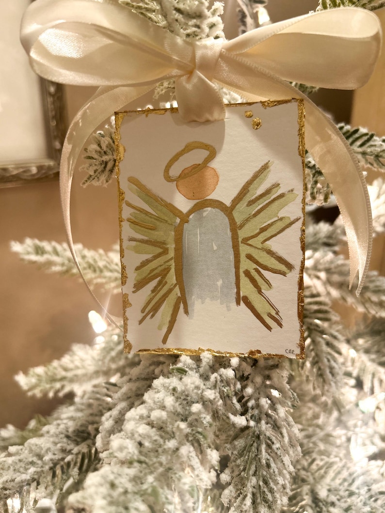 May include: A rectangular paper ornament featuring a hand-painted angel with a gold halo, light blue body, and gold-toned wings. The edges are accented with gold leaf, and the piece is suspended by a cream-colored satin ribbon bow.