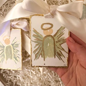 May include: Rectangular white ornaments featuring a hand-painted angel with a sage green body, gold halo, and gold wings. Each ornament has gold-leafed edges and is tied with a cream-colored satin ribbon. The artist's initials are visible in the corner.