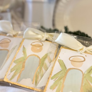 May include: Rectangular ornaments featuring a painted angel with a pale blue body, light green wings, and a gold halo. Each ornament has a gold-painted edge and is tied with a cream-colored satin ribbon, individually packaged in clear plastic sleeves.