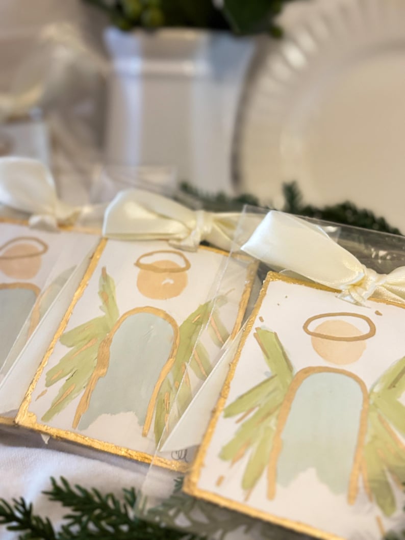 May include: Rectangular ornaments featuring a painted angel with a pale blue body, light green wings, and a gold halo. Each ornament has a gold-painted edge and is tied with a cream-colored satin ribbon, individually packaged in clear plastic sleeves.