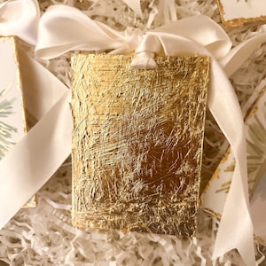 May include: A rectangular tag with a textured gold leaf finish on the front surface. The tag features a hole at the top center, threaded with a smooth, off-white satin ribbon tied into a bow. The gold surface displays visible brush strokes.
