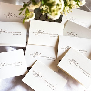 May include: A collection of ivory-colored cards with the text "With God, All Things Are Possible. Matthew 19:26" printed in a dark gray script. The cards are arranged on a light-colored surface, with a few flowers in the background.