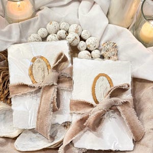 May include: Two white art prints with a gold oval design and a beige ribbon. The prints are wrapped in clear plastic. The word "Mother" is written on the bottom of one print. The background includes white fabric, decorative balls, and candles.