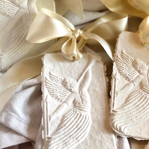 May include: Off-white rectangular clay ornaments featuring a raised relief of an angel playing a trumpet. Each ornament has a deckled edge and is tied with a smooth, cream-colored satin ribbon. The surface texture is matte and slightly porous.