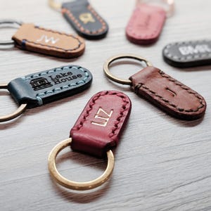 Può includere: Una collezione di sei portachiavi in ​​pelle con diversi design e incisioni. I portachiavi sono realizzati in pelle marrone, rossa, blu e nera. Le incisioni includono iniziali, nomi e la frase "Lake House".