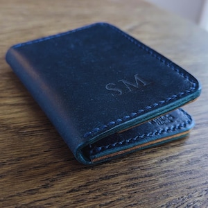 May include: A navy blue leather wallet with the initials "SM" embossed on the front. The wallet has a stitched border and a contrasting green edge. The wallet is closed and resting on a wooden surface.