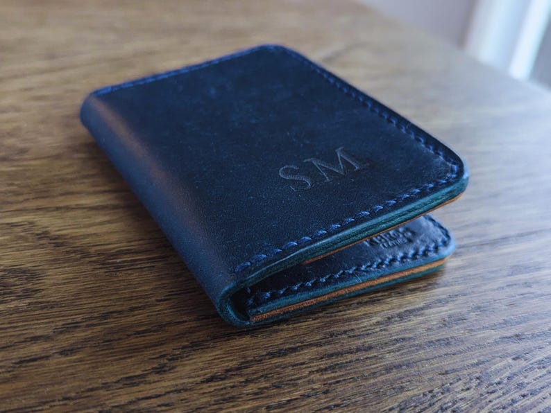 May include: A navy blue leather wallet with the initials "SM" embossed on the front. The wallet has a stitched border and a contrasting green edge. The wallet is closed and resting on a wooden surface.