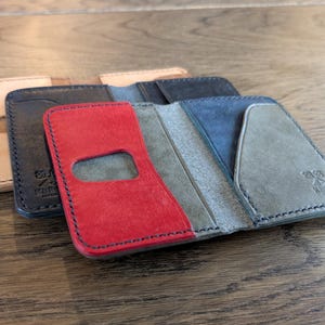 May include: A collection of handcrafted leather wallets in various colors. The wallets are open, showcasing their interior design. Colors include tan, black, red, blue, and gray. Each wallet has a unique design and stitching.