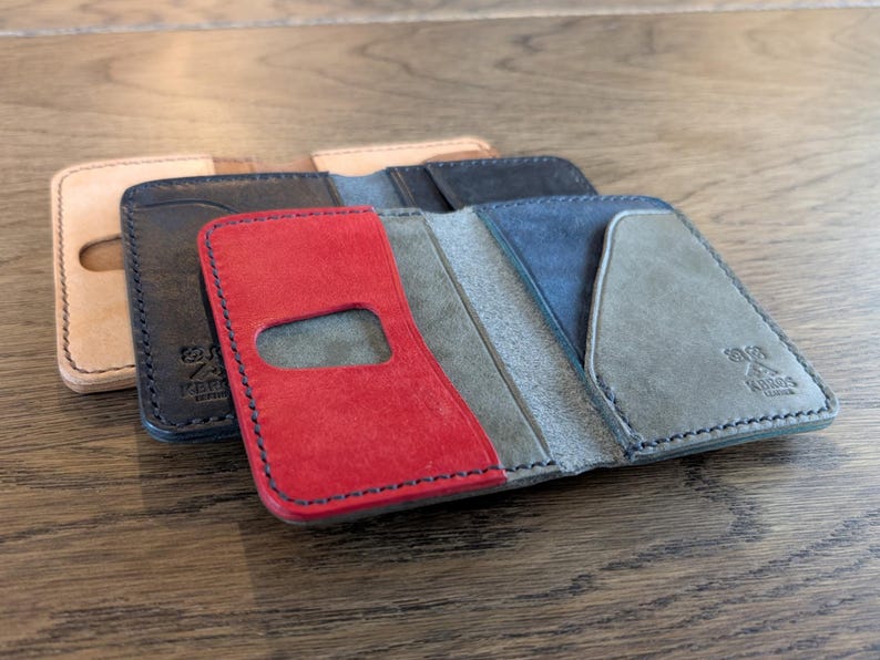 May include: A collection of handcrafted leather wallets in various colors. The wallets are open, showcasing their interior design. Colors include tan, black, red, blue, and gray. Each wallet has a unique design and stitching.