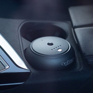 Puede incluir: Un ambientador de coche negro con el texto "OmBalance" y "Travel Essential" en él. El ambientador está colocado en un portavasos de un coche.