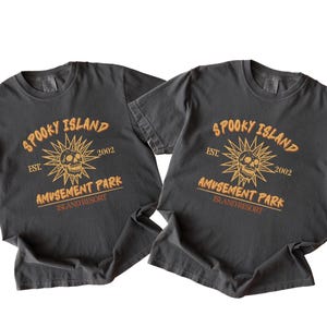 Peut inclure: Deux t-shirts gris foncé avec le texte "SPOOKY ISLAND AMUSEMENT PARK ISLAND RESORT" en orange. Le motif comprend un crâne dans un éclat de soleil et l'année 2002.
