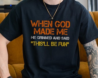 May include: Black t-shirt with the text "WHEN GOD MADE ME HE GRINNED AND SAID 'THIS'LL BE FUN'" in orange and yellow lettering. The shirt is a crew neck style.