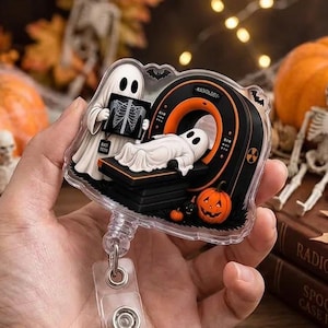 May include: A clear acrylic badge reel featuring a cartoon ghost technician holding an X-ray film and another ghost lying inside a black and orange MRI machine. The design includes a small orange jack-o'-lantern and a radiation symbol.