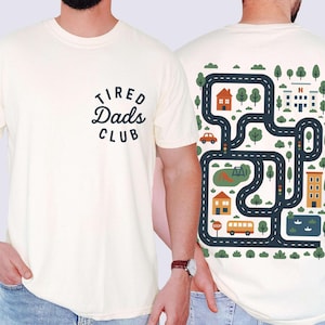 Może przedstawiać: Kremowa koszulka z napisem "Tired Dads Club" z przodu. Tył przedstawia wz&oacute;r mapy drogowej z domami, drzewami, autobusem i samochodem. Projekt jest w odcieniach niebieskiego, pomarańczowego i zielonego.