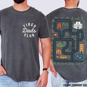 May include: Dark grey t-shirt with the text "TIRED Dads CLUB" on the front. The back features a colorful road map design with houses, trees, a hospital, and vehicles. The shirt is a Comfort Colors Pepper.