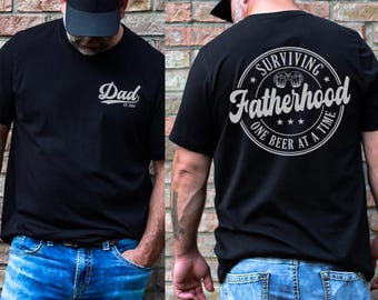 Può includere: T-shirt nera con la parola "Dad" in corsivo sul davanti, con "Est. 2024" sotto. Il retro della maglietta presenta un design circolare con le parole "Surviving Fatherhood One Beer at a Time."