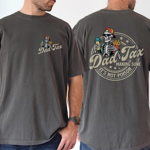 May include: Dark gray t-shirt with a graphic on the front and back. The front graphic says "Dad Tax" with a skeleton holding a drink and fries. The back graphic is a circular design with a skeleton, the words "Dad Tax", and "Making sure it's not poison."