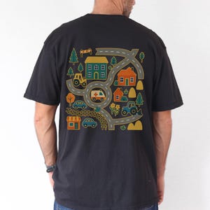 May include: Black t-shirt featuring a colorful graphic of a road map with houses, trees, cars, a tractor, and an ambulance. The design is in shades of yellow, green, orange, and blue. The shirt is a casual style.