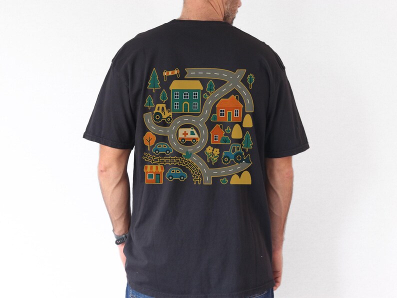 May include: Black t-shirt featuring a colorful graphic of a road map with houses, trees, cars, a tractor, and an ambulance. The design is in shades of yellow, green, orange, and blue. The shirt is a casual style.