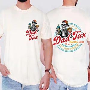 May include: Cream-colored t-shirt featuring a skeleton wearing a cap and sunglasses, holding a drink and fries. The text "Dad Tax" is in red script. The back of the shirt has the same graphic inside a blue circle with the text "Making Sure It's Not Poison."