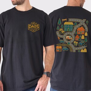 May include: Black t-shirt with a gold and white shield logo that reads "Tired Dads Club." The back of the shirt features a colorful graphic of a road map with houses, trees, and vehicles. The shirt is a casual, short-sleeved style.